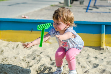 Little charming girl baby playing on the playground in the sandbox sand mound in the bucket with a shovel and rake