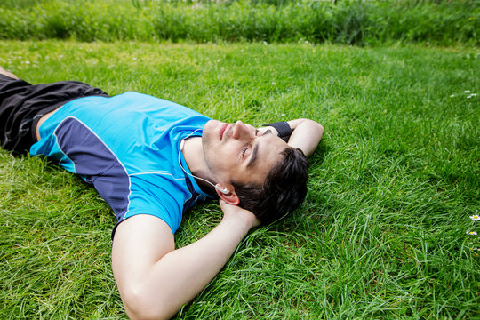 Sport fitness man relaxing listening to music after training outdoor in a city park . Young male athlete resting relaxing lying on grass after running and training exercise outside in summer.