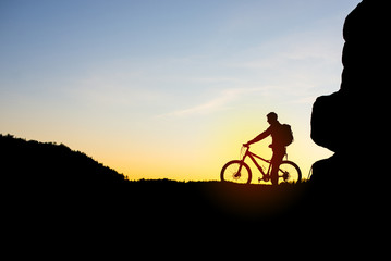 Silhouette of Cyclist with Bike on the Rock at Sunset.