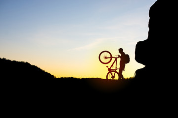 Silhouette of Cyclist with Bike on the Rock at Sunset.