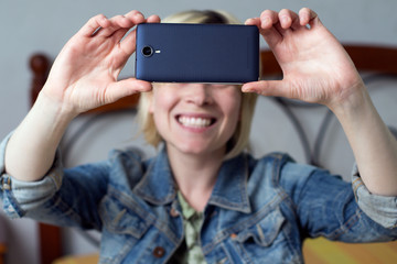 Blonde woman holding a mobile, smiling and  making a selfie in the room
