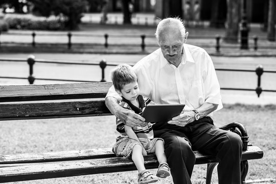 Grandchild Teaching To His Grandfather To Use Tablet On A Bench. Black And White Photography