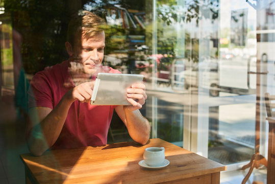 A young guy sitting at a table in a cafe and holding a tablet and clicks on the screen