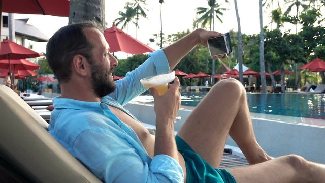 Young man on sunbed drinking cocktail and taking selfie photo with cellphone 
