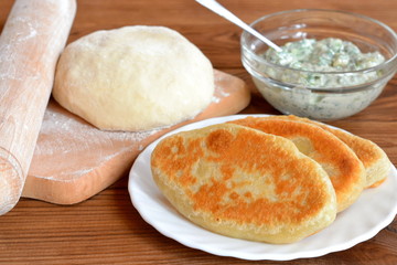 Fried pies on a plate. Dough and rolling pin on a cutting board. Cheese filling in a glass bowl. Wooden background. Home simply delicious food. Home cooking. Ingredients for making homemade pies