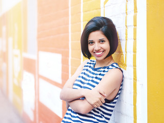 Portrait of beautiful smiling young hipster latin hispanic girl woman with short hair bob, in blue white striped tshirt, leaning on brick wall in city looking in camera, lifestyle fashion concept