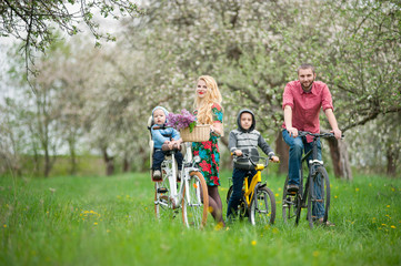 Happy family on a bicycles in the spring garden. Father and son on bikes, mom holding bike and baby sitting in bicycle chair, in the basket lay a bouquet of lilacs