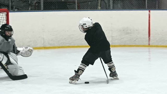 Little hockey player skating towards the opponent goalie, pulling stick between his legs and pushing puck into the net