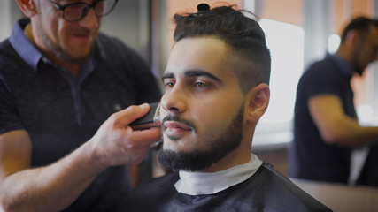 Young, brave hipster in your favorite barber shop. Master engaged in his beard, uses a trimmer to...