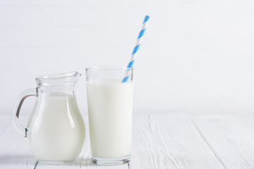 Glass of milk with stripped blue paper straw and jug of milk on white wooden table