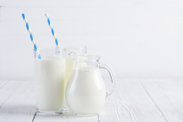 Glass of milk with stripped blue paper straw and jug of milk on white wooden table still life