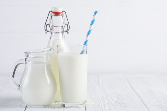 Glass of milk with stripped blue paper straw and jug with bottle of milk on white wooden table