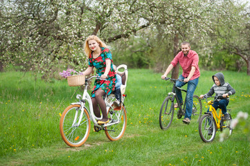 Young happy family with two kids riding bicycles in spring garden, little baby in bicycle chair, in the basket lay a bouquet of lilacs