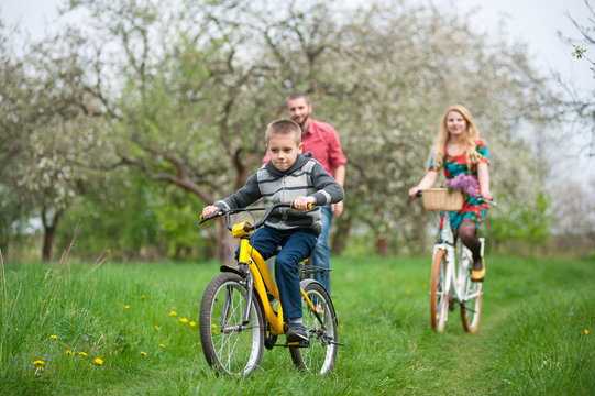 Happy boy on a bicycle in front of and behind its parent in the spring blooming garden. Family having fun against the fresh greenery. Front view
