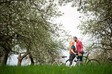 Loving young couple with bikes kissing against the background of blooming trees and fresh greenery in spring garden. Couple together enjoying romantic holidays. Side view