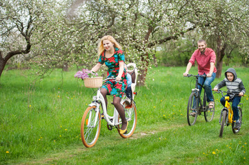 Young family with two kids riding bicycle in spring garden, little baby in bicycle chair, in the basket lay a bouquet of lilacs, against the background of blooming fresh greenery