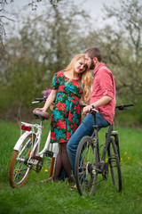 Obraz premium Loving beautiful couple with bicycles against the background of trees and fresh greenery in spring garden. Woman looking to the camera. Couple together enjoying romantic holidays