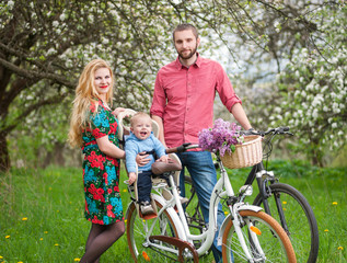 Happy family on a bicycles in the spring garden. Mother holding bike and baby sitting in bicycle chair. Against the background of blooming fresh greenery