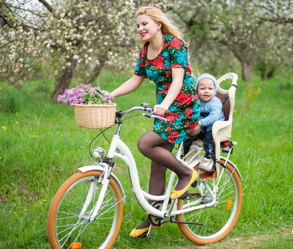 Blonde female with long hair in dress riding city bicycle with baby in bicycle chair, in the basket lay a bouquet of lilacs, against the background of blooming fresh greenery in spring garden