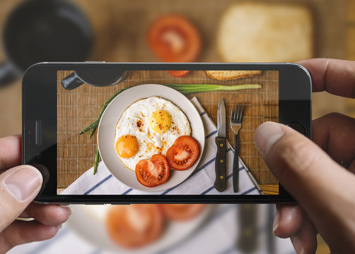 Taking picture of fried eggs and tomato with mobile phone. Phone in male hands.On the plate there is 2 fried eggs, tomato, fork and knife. Vintage style.