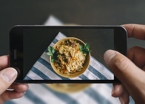 Taking picture of fried rice bowl with mobile phone. Phone in male hands.On the plate there is fried rice with pork. Plate is on a black aged wooden table. This fried rice called pilaf. Vintage style.