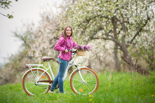 Smiling brunette woman wearing purple jacket and jeans with a vintage white bicycle and lilac flowers basket, against the background of blooming trees, dandelions and fresh greenery in spring garden