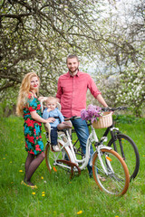 Fototapeta na wymiar Beautiful family on a bicycles in the spring garden. Mother holding her bike and baby sitting in bicycle chair, against the background of blooming fresh greenery