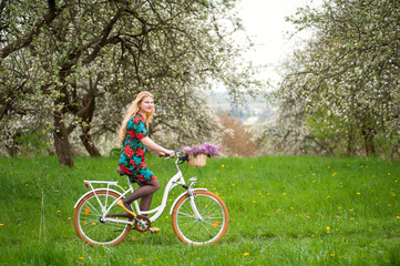 Happy woman with long blond hair wearing flowered dress and yellow shoes riding a vintage white bicycle. Blooming trees, dandelions and fresh greenery in spring garden on the background