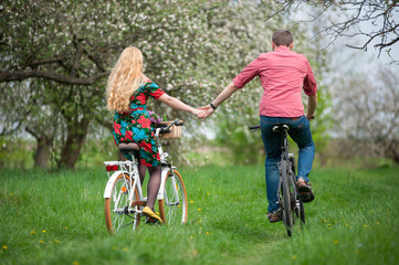 Happy couple riding bicycles in the spring garden back to camera and holding hands. Female with long blond hair wearing flowered dress and man in a red shirt