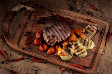 Overhead view of grilled marble beef steak with roasted vegetables: sliced vegetable marrow, bell pepper and cherry tomatoes on wooden board on rustic wooden counter