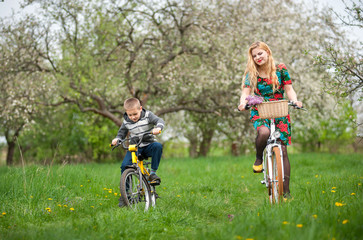Mother with son riding bicycles against the background of blooming fresh greenery in spring garden. Female wearing flowered dress and yellow shoes