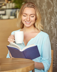 Beautiful young woman sitting at the table, reading a book and holding a cup of drink in the cafe.