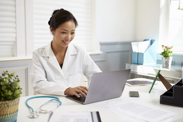 Female Doctor Sitting At Desk Working At Laptop In Office - Powered by Adobe
