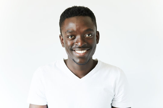 Portrait of handsome young African black male wearing white T-shirt looking and smiling at the camera with cheerful and happy expression, relaxing indoor against white studio wall background
