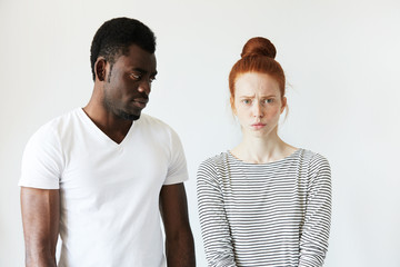 Extraordinary young interracial couple. Beautiful Caucasian woman looking at the camera with offended expression standing next to her boyfriend looking at her with angry and disappointed face