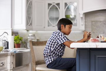 Young Asian Boy Playing Game On Mobile Device In Kitchen
