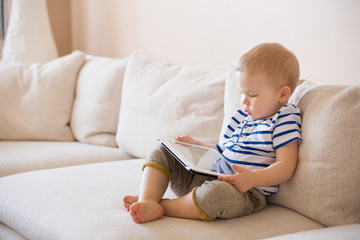 Adorable blond toddler boy laying on the sofa and playing with tablet pc at home, indoors. Child with tablet computer.
