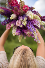 Fototapeta premium Back view on young girl holding a bunch of beautiful lupine flowers. Purple and pink lupin bouquet. Womanl with bouquet of wild lupin flowers on sunny summer field. Sky background.