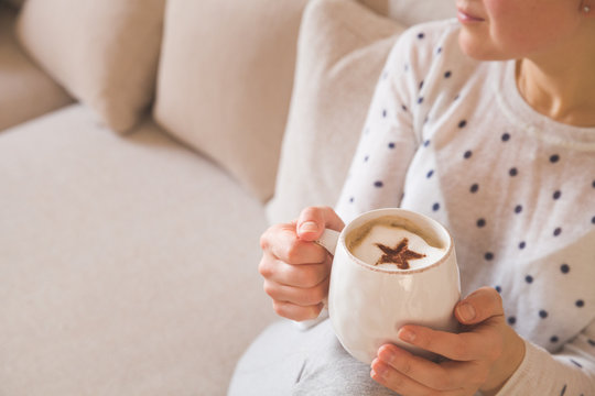 Girl with coffee mug sitting on the sofa indoors. Woman drinking a cup of  coffee or tea sitting cozy at home. Relax and rest. Cappuccino with cinnamon star.