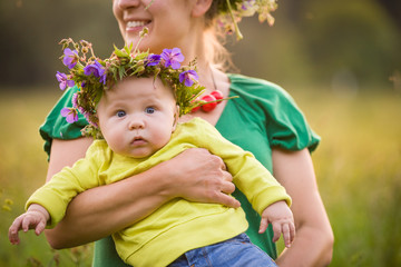 Happy mother holding her adorable little baby boy on a summer meadow. Mother with kid in flower wreath. Family together, parent with little child. Woman hugging baby on sunny day.