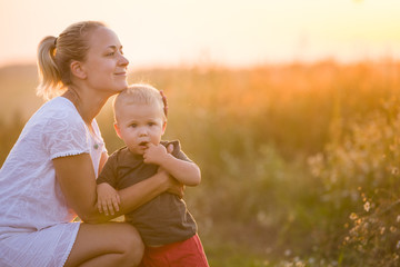 Young beautiful mother hugging her little toddler son on a sunset in a wild flowers field. Happy...