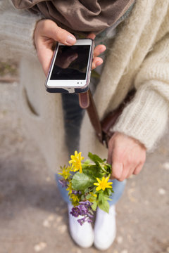 woman holding a beautiful bunch of wild spring flowers and taking photo of it with her smartphone. Bouquet of yellow and blue flowers in hands. Making picture by the telephone.