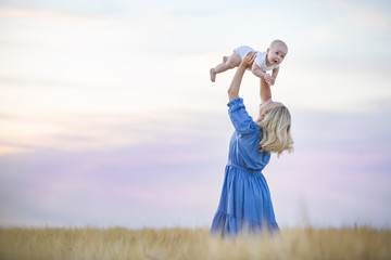 Beautiful young mother holding her little baby boy at sunset. Mother and baby in the wheat field on a sunny summer day. Happy woman with adorable child. Motherhood and childhood concept.