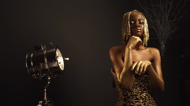 Creative surreal portrait of sexy african american female model with glossy golden makeup and headwear posing to the camera. Bronze bodypaint, black studio background. Vogue concept