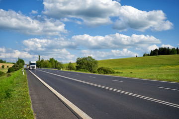 Asphalt road with trucks arriving from far away in the countryside. Sunny day with blue skies and white clouds over green meadows.