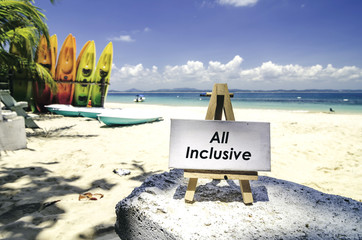 conceptual image,white canvas frame and wooden easel with word ALL INCLUSIVE. Tropical beach  at sunny day ,white sand,colorful kayaks and clear blue water background.