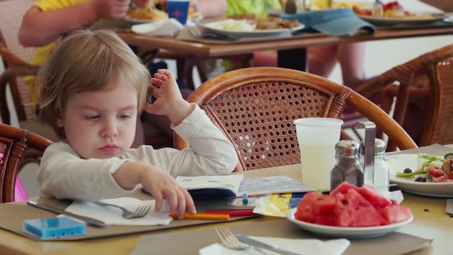 Little child seating at the restaurant, refusing eat watermelon, drawing something.