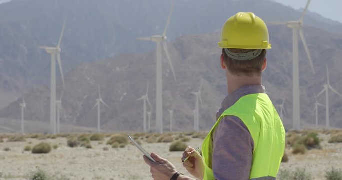 Technician in hard hat and wearing yellow high-visibility vest, at wind farm, using a tablet computer and stylus. Walks off frame at end. Medium close up, originally recorded in 4K.