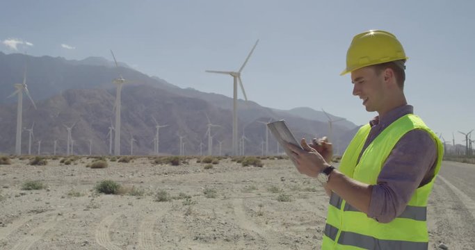 Technician in hard hat and wearing yellow high-visibility vest, at wind farm, using a tablet computer and stylus. Holds finger up to check wind direction.  Medium shot, originally recorded in 4K.