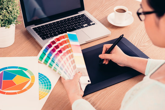 image of a graphic designer using graphic tablet in her work on the foreground and working with color samples for selection. A concept of modern digital workplace